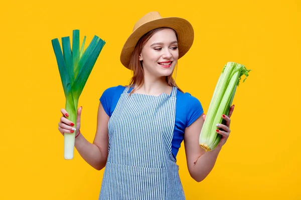 Heureuse jeune femme caucasienne agricultrice tenant poireau et céleri légumes dans ses mains sur fond jaune Images De Stock Libres De Droits
