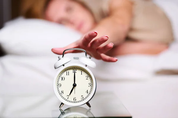 Slaperige jonge blanke vrouw die haar hand uitstrekt tot het alarm afgaat om het uit te zetten. Vroeg wakker worden, niet genoeg slapen, werkconcept krijgen. — Stockfoto