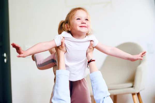 Joven madre juega con su hija preescolar en casa y la cría en sus brazos, el niño vuela como un avión — Foto de Stock