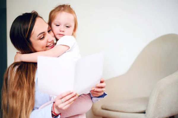 Feliz Día de las Madres. Feliz mamá sonriente con su pequeña hija leyendo la tarjeta de vacaciones mientras está sentada en el suelo — Foto de Stock