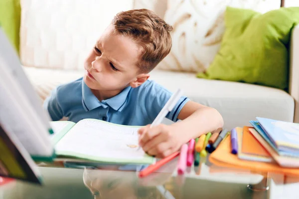 Думая о ребенке скучно, маленький мальчик пишет внимательно сидя за столом и делать домашнее задание — стоковое фото