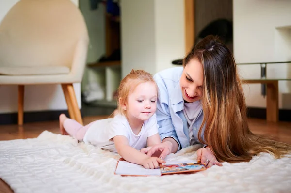 Madre joven con su linda hija pequeña está leyendo un libro y mirando los dibujos en el libro mientras está acostada en el suelo. La madre enseña al niño. La relación entre padres e hijos — Foto de Stock