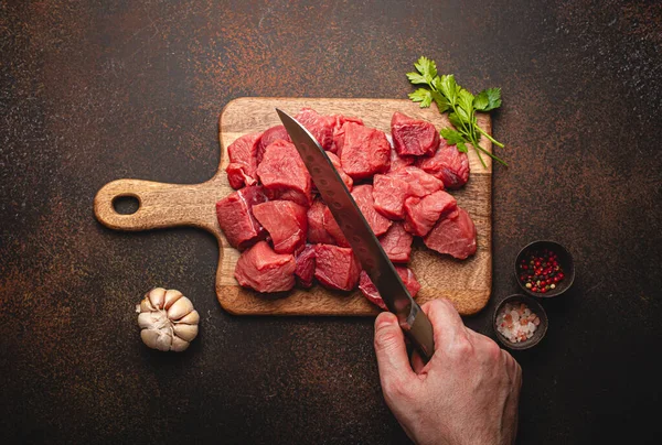Manlig hand hugga i kuber rått nötkött kött på träskiva — Stockfoto