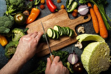 Man hands chopping cucumber on wooden cutting board with knife and assorted fresh vegetables