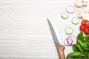 Food cooking background: fresh ingredients with kitchen knife, basil, cherry tomatoes, mushrooms, cut cucumber and onion on white wooden background top view. Meal preparing template, space for text