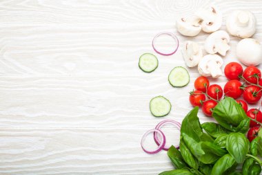 Food cooking background: fresh ingredients basil, cherry tomatoes, mushrooms, cut cucumber and onion on white wooden background top view. Meal preparing template, space for text