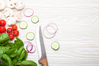 Food cooking background: fresh ingredients with kitchen knife, basil, cherry tomatoes, mushrooms, cut cucumber and onion on white wooden background top view. Meal preparing template, space for text