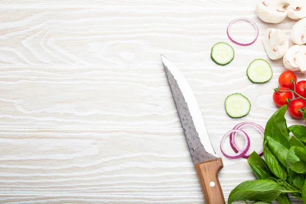 Food cooking background: fresh ingredients with kitchen knife, basil, cherry tomatoes, mushrooms, cut cucumber and onion on white wooden background top view. Meal preparing template, space for text