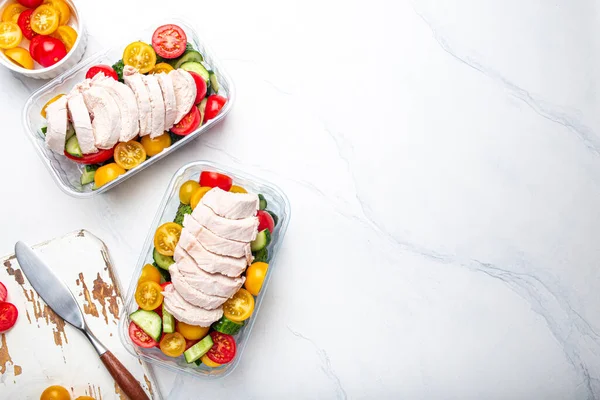 Healthy meal prep for lunch, diet boiled chicken fillet with vegetables salad in lunch boxes containers top view on white marble kitchen table background with knife and cutting board. Space for text