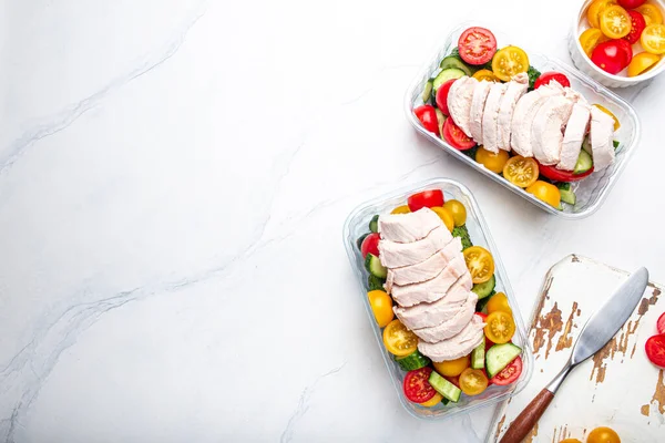 Healthy meal prep for lunch, diet boiled chicken fillet with vegetables salad in lunch boxes containers top view on white marble kitchen table background with knife and cutting board. Space for text