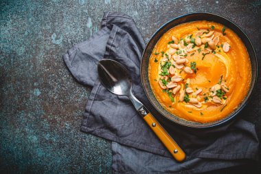 Koyu taştan arka planda yer fıstıklarıyla süslenmiş, yukarıdan süslenmiş balkabağı kremalı bir seramik kâse. Sonbahar sıcacık akşam yemeği konsepti, metin için boşluk