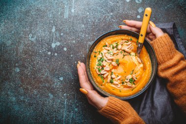 Sarı örülü süveter giyen kadın elleri, kabak kremalı bir kaseyi arka planda yer fıstığıyla süslenmiş bir kaşık üzerinde tutuyordu. Sonbahar sıcacık akşam yemeği konsepti, metin için boşluk.