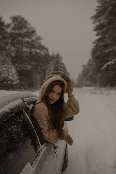 若い女の子が車の窓から腰に手を上げた 森の中の冬の道に若い女の子が車の窓から出てきた 縦の写真 セレクティブフォーカス — ストック写真