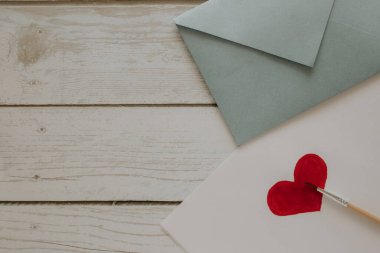 Mavi bir zarf ahşap bir arka planda yatar, yanında çizilmiş bir kalp ve fırça olan bir yaprak yatar. Metin için yer.