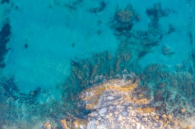 Turkuaz deniz suyunda kayalara çarpan dalgaların yukarıdan aşağı görüntüsü.