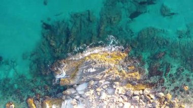 Turkuaz deniz suyunda kayalara çarpan dalgaların havadan aşağı görüntüsü.
