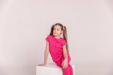 Pembe tişörtlü ve pantolonlu, beyaz izole bir arka planı olan güzel bir kız. Beyaz bir küpün üzerinde oturan çocuk. Kuyruklu kız. Boşluğu kopyala