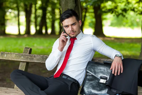 Young business man talking on smart phone in city park sitting on the bench during the break — Stock Photo, Image