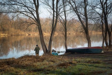 Kamuflaj pantolonu ve kapüşonlu ceketli bir adam sonbahar sabahı nehirde balık tutuyor..