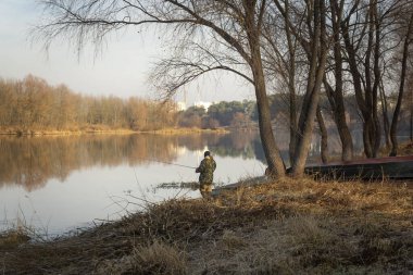 Kamuflaj pantolonu ve kapüşonlu ceketli bir adam sonbahar sabahı nehirde balık tutuyor..