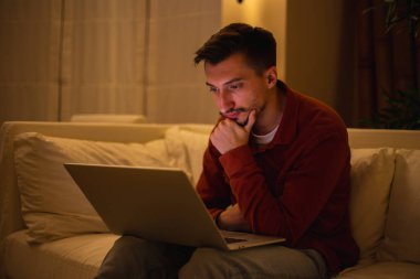 Kırmızı tişörtlü sakallı genç bir adam dizüstü bilgisayarla çalışıyor ve akşamları evdeki kanepede oturuyor..