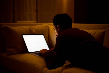 Kırmızı tişörtlü sakallı genç bir adam dizüstü bilgisayarla çalışıyor ve akşamları evdeki kanepede oturuyor..