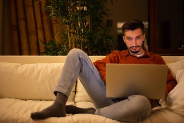 Kırmızı tişörtlü sakallı genç bir adam dizüstü bilgisayarla çalışıyor ve akşamları evdeki kanepede oturuyor..