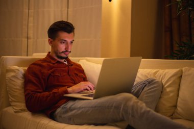 Kırmızı tişörtlü sakallı genç bir adam dizüstü bilgisayarla çalışıyor ve akşamları evdeki kanepede oturuyor..