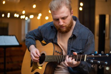 A young guy with a beard plays an acoustic guitar in a room with warm lighting