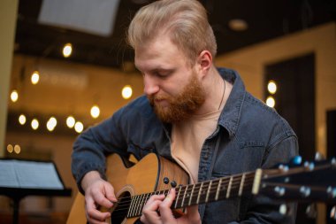 A young guy with a beard plays an acoustic guitar in a room with warm lighting