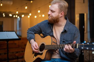 A young guy with a beard plays an acoustic guitar in a room with warm lighting