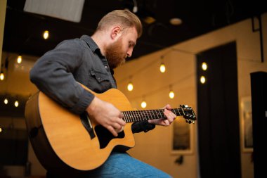 A young guy with a beard plays an acoustic guitar in a room with warm lighting