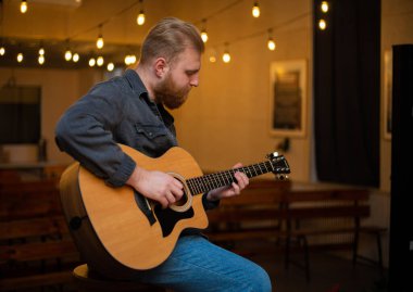 A young guy with a beard plays an acoustic guitar in a room with warm lighting