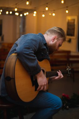 A young guy with a beard plays an acoustic guitar in a room with warm lighting