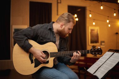 A young guy with a beard plays an acoustic guitar in a room with warm lighting