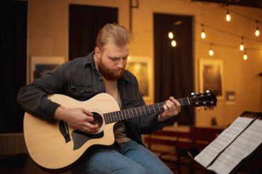 A young guy with a beard plays an acoustic guitar in a room with warm lighting