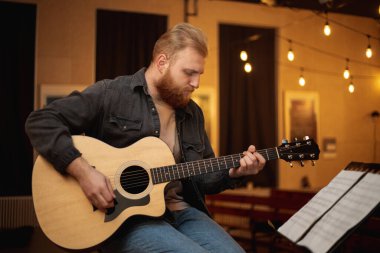 A young guy with a beard plays an acoustic guitar in a room with warm lighting