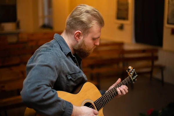 A young guy with a beard plays an acoustic guitar in a room with warm lighting — 图库照片