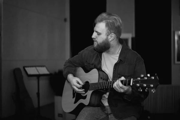 A young guy with a beard plays an acoustic guitar in a room with warm lighting — 图库照片