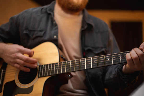 Молодий хлопець з бородою грає на акустичній гітарі в кімнаті з теплим освітленням — стокове фото