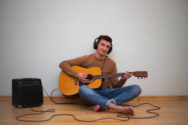 Genç bir adam arka planda duvarla gitar çalıyor.