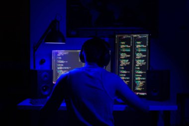 Bir adam geceleri bir masada bilgisayarın başında oturuyor. Mavi ışık ve programlarla.