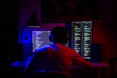 Bir adam geceleri bir masada bilgisayarın başında oturuyor. Mavi ışık ve programlarla.