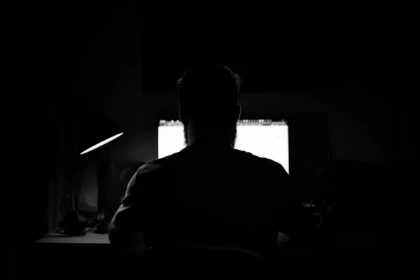 A man sits at a computer in a room at a table at nigh programming black and white — Stock Photo, Image
