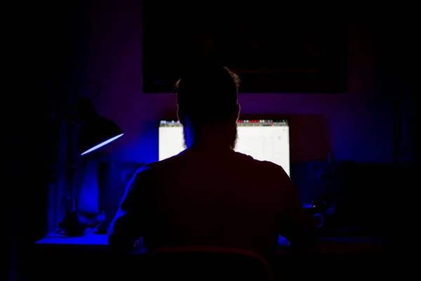 Bir adam geceleri bir masada bilgisayarın başında oturuyor. Mavi ışık ve programlarla.