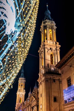 Basilica del Pilar 'ın kuleleri, İspanya' nın Zaragoza kentindeki Plaza del Pilar 'da gece belediye binasını aydınlatan ışık topunun ve Noel ışıklarının aydınlatılmasıyla odaklanmış durumda..