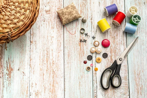 top view of sewing items such as scissors, a thimble, various buttons and spools of colored thread a wicker sewing box on a vintage wooden table