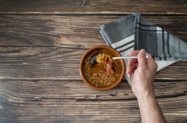 Kırsal ahşap bir masada, üzerinde yazı için yeri olan kil bir çanaktan çıkan mercimeği kaşıkla tutan bir elin üst görüntüsü. Sağlıklı ve geleneksel İspanyol mutfağı