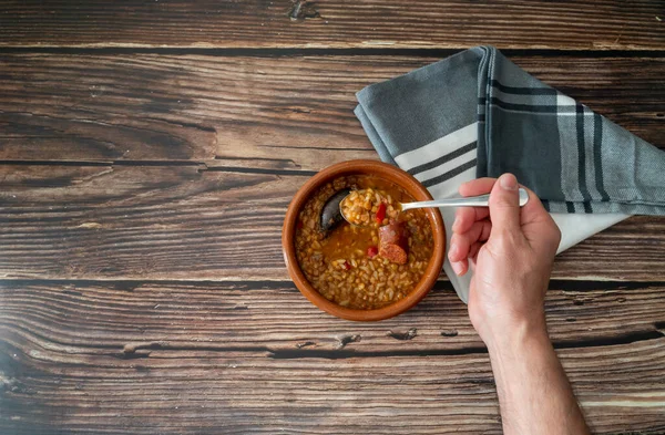 Kırsal ahşap bir masada, üzerinde yazı için yeri olan kil bir çanaktan çıkan mercimeği kaşıkla tutan bir elin üst görüntüsü. Sağlıklı ve geleneksel İspanyol mutfağı