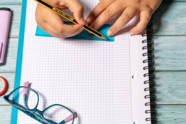 top shot of a girl's hands using a pencil and ruler on a grid notebook with modern glasses on a blue wooden table. Material concept for the new school year